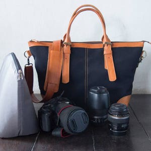 May include: A black canvas camera bag with brown leather accents and straps. The bag is open and a camera lens and a camera body are visible inside. A gray camera bag is also in the image.