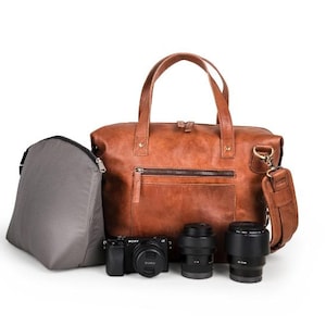 Peut inclure: Un sac à dos en cuir marron pour appareil photo avec une poignée supérieure et une bandoulière réglable. Le sac est ouvert et contient trois objectifs d'appareil photo et un appareil photo.