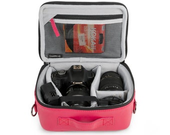 May include: A vibrant pink camera bag, open to show a Canon DSLR camera, lenses, and accessories. The interior is grey with padded dividers. A mesh pocket holds extra items. The bag has a top handle and a zip closure.