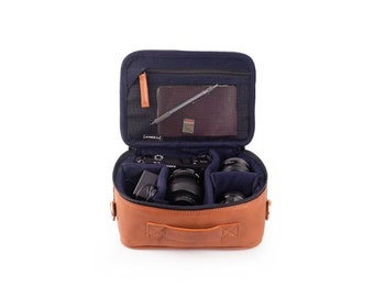 May include: A brown leather camera bag with a blue interior. The bag is open and contains a camera, lenses, and a charger.