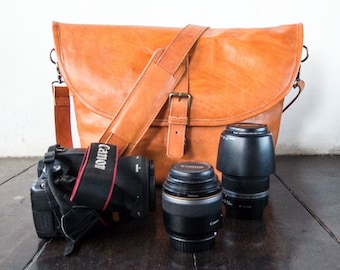 May include: A brown leather camera bag with a shoulder strap, a black camera with a lens, and two additional lenses. The camera bag is on a wooden surface.