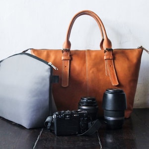 Könnte beinhalten: Eine Sammlung von Fotoausrüstung, darunter eine braune Ledertasche, eine graue Kameratasche, eine schwarze Kamera und zwei Objektive. Die Tragetasche hat zwei Griffe und Schnallen.