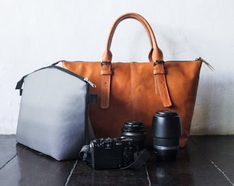 May include: A collection of photography equipment, including a brown leather tote bag, a gray camera bag, a black camera, and two camera lenses. The tote bag has two handles and a buckle detail.