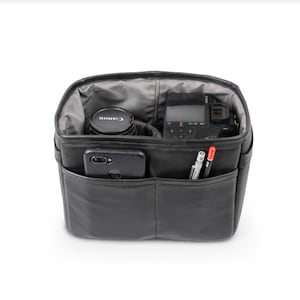 Puede incluir: Bolsa de cámara negra con múltiples bolsillos y compartimentos. La bolsa está abierta y contiene una cámara, un objetivo, un teléfono y bolígrafos.