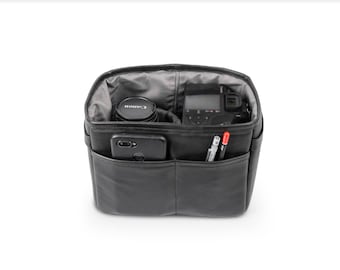 May include: Black camera bag with multiple pockets and compartments. The bag is open and contains a camera, lens, phone, and pens.