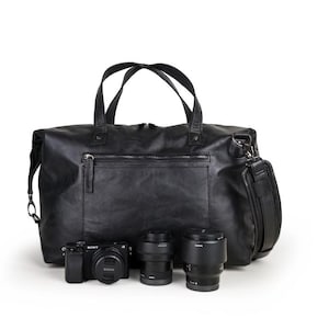 Puede incluir: Una bolsa de cámara de cuero negro con asa superior y correa para el hombro. La bolsa está abierta y contiene una cámara negra y dos lentes de cámara negros.