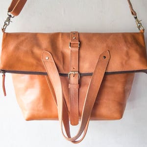 May include: A large, tan leather tote bag with a zipper closure and a buckle strap. The bag has two long straps that can be used to carry it on the shoulder or by hand.