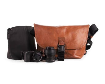 May include: A brown leather camera bag with a black strap and a black camera and lens inside. The bag is sitting on a white surface.