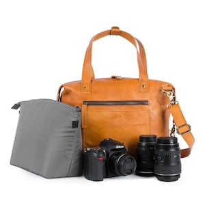Puede incluir: Una bolsa de cámara de cuero marrón con un inserto acolchado gris. La bolsa tiene un asa superior y una correa para el hombro. La bolsa está abierta y contiene una cámara negra y dos lentes de cámara negras.