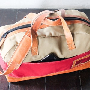 Pu&ograve; includere: Una borsa da viaggio in tela beige e rossa con cinturini e rifiniture in pelle marrone. La borsa ha una chiusura a zip e una piccola tasca sul davanti.