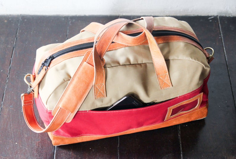 Pu&ograve; includere: Una borsa da viaggio in tela beige e rossa con cinturini e rifiniture in pelle marrone. La borsa ha una chiusura a zip e una piccola tasca sul davanti.
