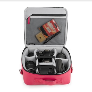 Peut inclure: Sac à dos rose pour appareil photo avec un appareil photo noir et un objectif à l'intérieur. Le sac a une poche zippée à l'avant et une poche en filet à l'intérieur. Le sac est ouvert et l'appareil photo est visible.