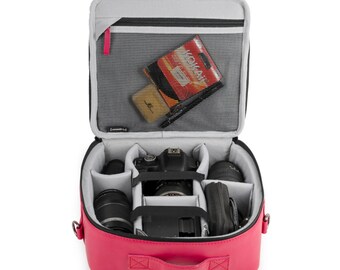 May include: Pink camera bag with a black camera and lens inside. The bag has a zipped pocket on the front and a mesh pocket on the inside. The bag is open and the camera is visible.