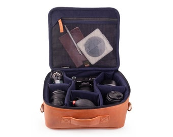 May include: A brown leather camera bag with a blue interior. The bag is open and contains a camera, lenses, and other accessories. The bag has a zipped pocket on the top and a handle on the front.