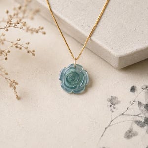 May include: A delicate gold chain necklace featuring a carved, translucent blue-green stone pendant shaped like a blooming rose. The pendant is suspended from a thin, polished gold-tone metal chain, showcasing a smooth, glossy finish.