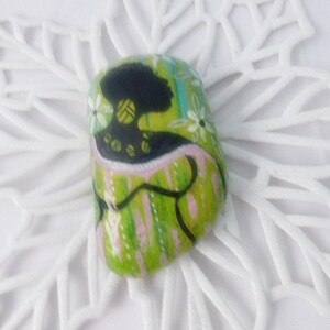 May include: A green, pink, and black painted rock with a stylized image of a woman with a green and black patterned dress. The rock is on a white background.