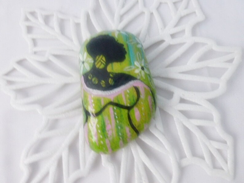 May include: A green, pink, and black painted rock with a stylized image of a woman with a green and black patterned dress. The rock is on a white background.