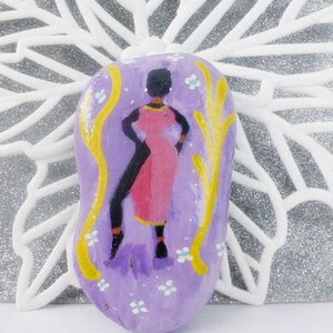 May include: Hand-painted stone featuring a stylised figure of a woman in a pink dress and yellow accents on a purple background with white flowers.