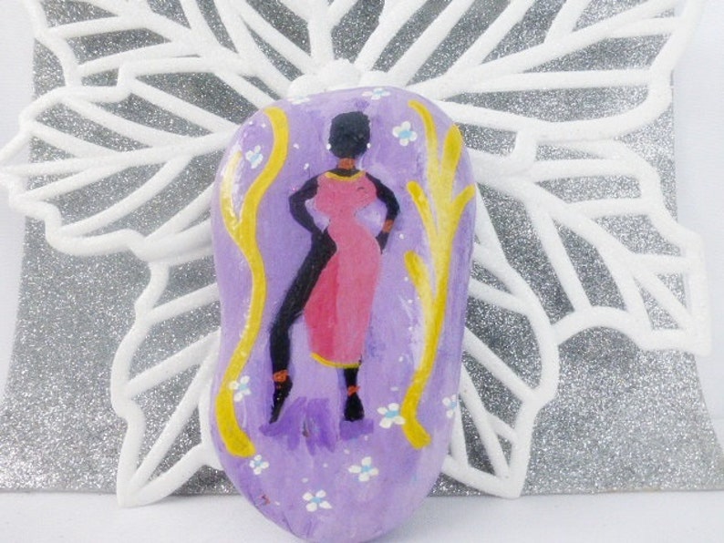 May include: Hand-painted stone featuring a stylised figure of a woman in a pink dress and yellow accents on a purple background with white flowers.