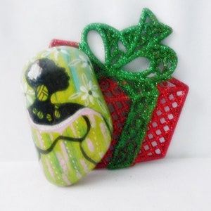 May include: A green and white painted ornament with a silhouette of a woman in a green dress. The ornament is decorated with white flowers and is sitting on top of a red and green glitter gift box.