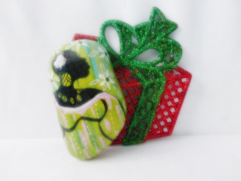 May include: A green and white painted ornament with a silhouette of a woman in a green dress. The ornament is decorated with white flowers and is sitting on top of a red and green glitter gift box.