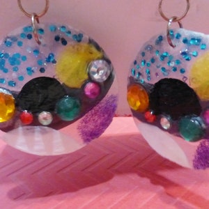 Può includere: Un paio di orecchini con una base in resina trasparente e decorazioni colorate. Gli orecchini presentano un cerchio nero con una varietà di strass colorati, glitter e peluria viola.