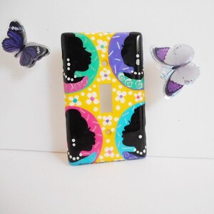 May include: A decorative light switch cover with a yellow background, featuring black silhouette profiles, and colorful accents of pink, teal, and purple. White flowers and butterflies add to the design.