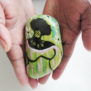 May include: A painted rock with a green, pink, and white background. A black silhouette of a woman with a yellow necklace is painted on the rock.