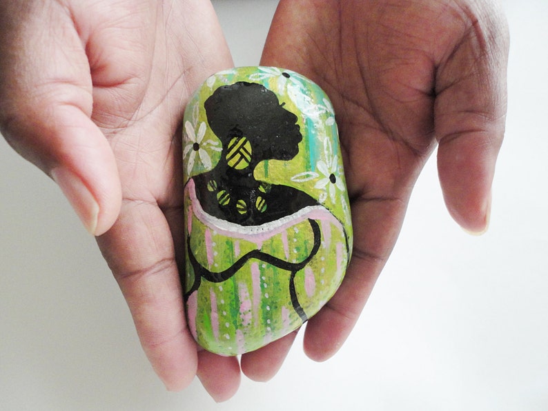May include: A painted rock with a green, pink, and white background. A black silhouette of a woman with a yellow necklace is painted on the rock.