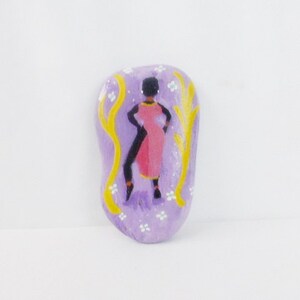 May include: A hand-painted stone with a purple background and white flowers. A black silhouette of a person in a pink dress stands in front of two yellow branches.