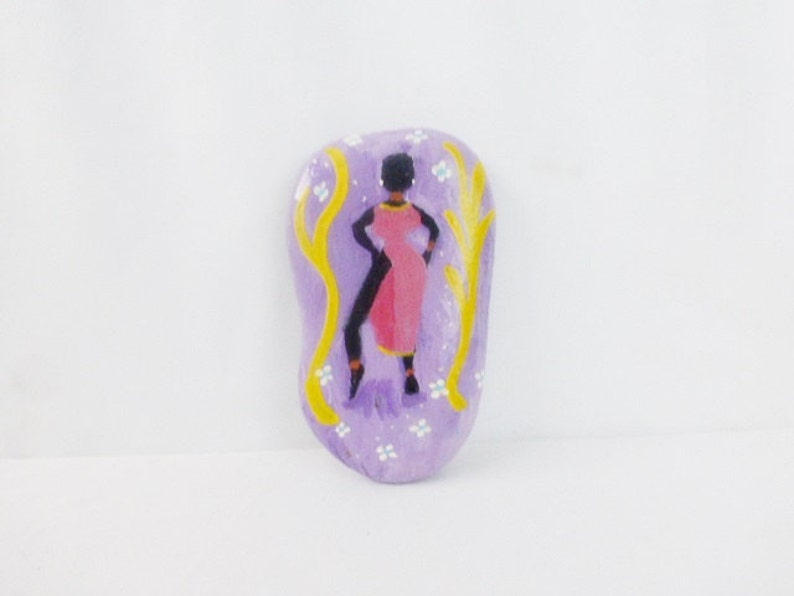 May include: A hand-painted stone with a purple background and white flowers. A black silhouette of a person in a pink dress stands in front of two yellow branches.
