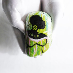 May include: A painted rock with a green, white, and pink background. A black silhouette of a woman with a floral design is painted on the rock.