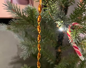 May include: A gold-colored, spiral-shaped Christmas tree ornament hanging from a green pine branch. The ornament has a twisted design and is suspended by a small loop. A candy cane is also visible.