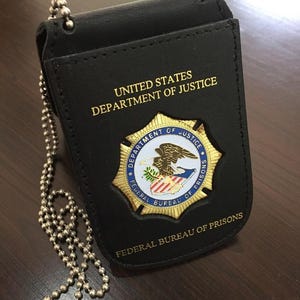 Puede incluir: Un porta-placas de cuero negro con una cadena de bolas plateada. La placa presenta el texto "UNITED STATES DEPARTMENT OF JUSTICE" en letras doradas. Un emblema dorado y azul dice "DEPARTMENT OF JUSTICE FEDERAL BUREAU OF PRISONS".