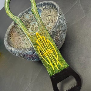 May include: A green and gold glitter bottle opener with a black opening. The opener has a yellow design on the handle. The opener is resting on a textured, gray bowl.