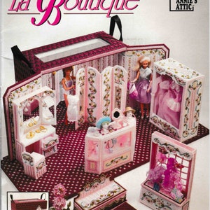 May include: A plastic canvas craft kit for a dollhouse boutique. The kit includes a pink and white dollhouse with a pink and white counter, a pink and white bench, a pink and white dressing room, and a pink and white display case. The kit also includes a pink and white doll, a pink and white dress, and a pink and white hat. The kit is from Annie's Attic.