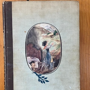 May include: A vintage book cover with a light blue background and a circular illustration of a woman in a dress standing in a mountainous landscape. The woman is looking up at the sky and her arms are outstretched. The illustration is surrounded by a dark brown oval frame and a green leafy branch.