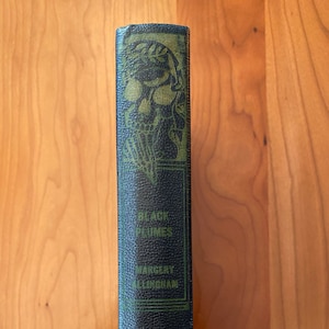 Puede incluir: Un libro negro con una encuadernación verde. La encuadernación tiene un diseño de calavera y huesos cruzados y el título "Black Plumes" de Margery Allingham. El libro está publicado por Doubleday Doran.