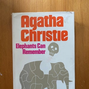 Könnte beinhalten: Ein Vintage-Taschenbuch mit dem Titel "Elephants Can Remember" von Agatha Christie. Das Cover ist weiß mit orangefarbenem und rosa Text, einem grauen Puzzle-Elefanten und einer Totenkopf-Grafik.
