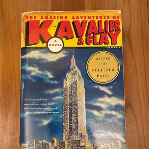 May include: A book cover with a red, white, and blue illustration of a cityscape at night. The title of the book is "The Amazing Adventures of Kavalier & Clay" and the author is Michael Chabon. The book is a novel and a winner of the Pulitzer Prize.