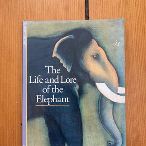 Könnte beinhalten: Ein Buchcover mit einer detaillierten Illustration eines Elefanten vor einem blauen und grünen Hintergrund. Der Titel des Buches lautet "The Life and Lore of the Elephant" von Robert Delort. Das Buch ist Teil der Reihe "Discoveries".