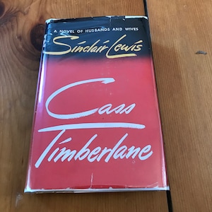 Może przedstawiać: Czerwona książka w twardej oprawie z tytułem "Cass Timberlane" w białym, ozdobnym pismem. Autor, Sinclair Lewis, jest nadrukowany czarnym, ozdobnym pismem nad tytułem. Okładka książki ma czarne tło z nadrukowanym białym tekstem "A Novel of Husbands and Wives".