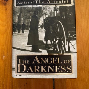 Op de afbeelding: Boekomslag voor "De Engel der Duisternis" van Caleb Carr, auteur van "De Alienist". De omslag toont een zwart-witfoto van een man in een lange jas die op straat staat voor een koets met paarden.