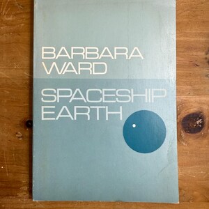 Może przedstawiać: Vintage okładka książki z tytułem "Spaceship Earth" autorstwa Barbary Ward. Okładka jest jasnoniebieska z białym tekstem i turkusowym okręgiem z białą kropką. Książka leży na drewnianej powierzchni.