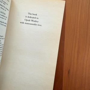 K&ouml;nnte beinhalten: Offene Buchseite mit dem Text: &bdquo;Dieses Buch ist Oprah Winfrey mit unermesslicher Liebe gewidmet.&ldquo; Die Seite ist cremefarben, der Text ist schwarz. Das Buch liegt auf einer Holzoberfl&auml;che.