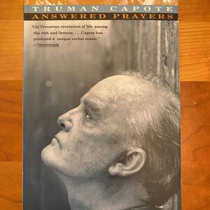 May include: A black and white photo of Truman Capote, the author of Answered Prayers, on the cover of a book. The quote "[A] Proustian revelation of life among the rich and famous.... Capote has produced a unique verbal music." - Newsweek is printed below the photo.