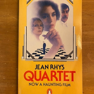 May include: A yellow paperback book with a black and white checkered pattern on the cover. The book title is "Quartet" by Jean Rhys. The book cover features a black and white photo of a woman with a man behind her. The book cover also includes the text "Now a Haunting Film" and a penguin logo.
