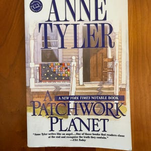 May include: A paperback copy of Anne Tyler's "A Patchwork Planet". The cover shows a house illustration with a patchwork quilt. The book is a New York Times bestseller. The author's name and title are in large blue letters.