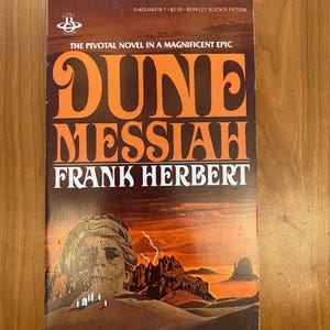 May include: A vintage paperback book titled "Dune Messiah" by Frank Herbert. The cover features orange and brown text with a desert landscape illustration. The book is part of the Berkley Science Fiction series.