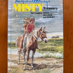 Puede incluir: Un libro de tapa dura titulado "Misty Treasury" de Marguerite Henry. La portada presenta una ilustración de dos niños montando a caballo cerca de una masa de agua. El título está en amarillo, con el nombre del autor en azul. El libro incluye "The Complete Misty, Sea Star and Stormy".
